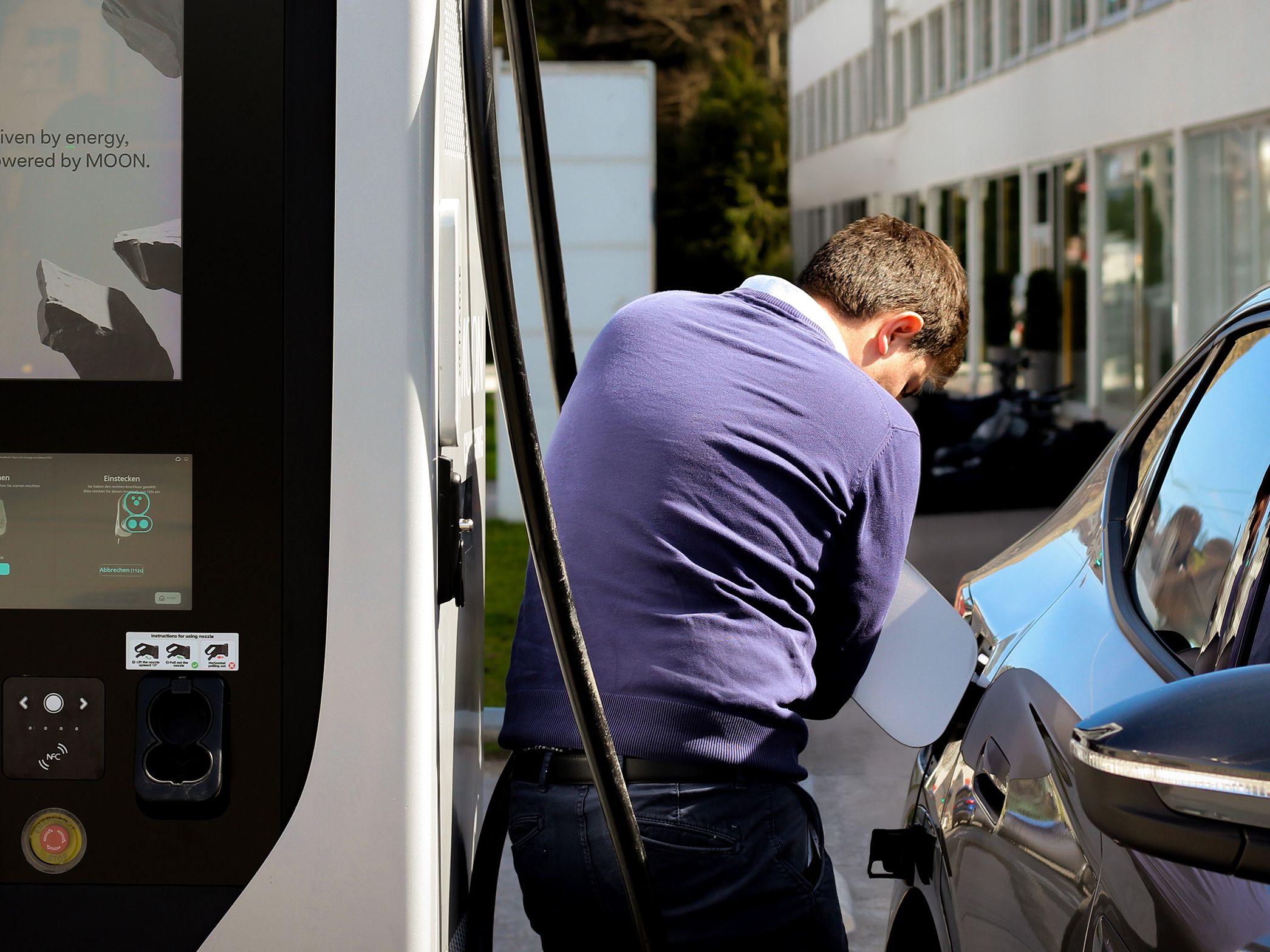 Moški odpira pokrov polnilnega priključka električnega vozila ob hitri polnilni postaji.