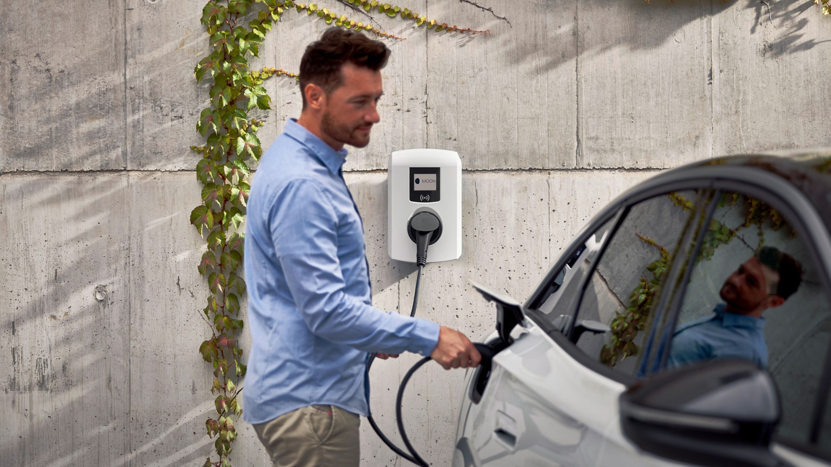 Moški pred stavbo priklaplja električni avto na stensko polnilnico.