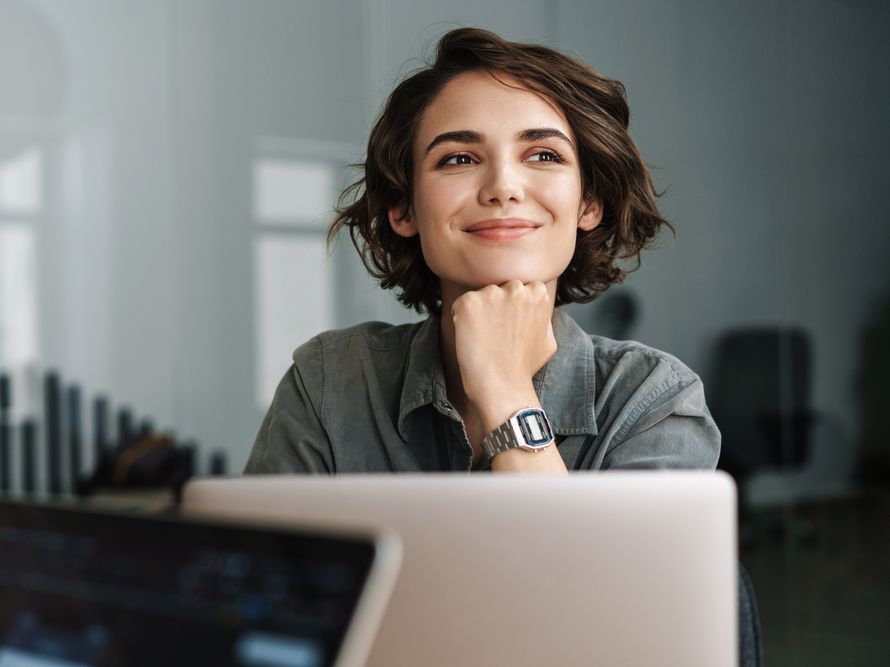 Ženska v pisarni sedi za prenosnim računalnikom in se zamišljeno nasmehne.