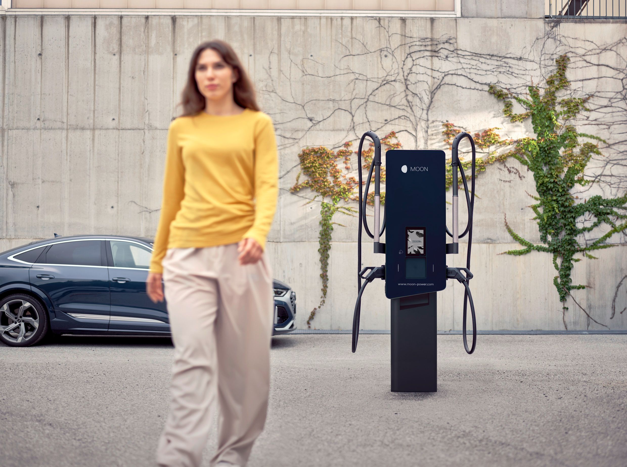 Ženska odhaja od MOON stenske polnilnice, ob kateri je parkiran električni avtomobil.