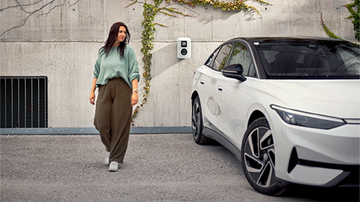 Ženska hodi ob električnem avtomobilu pred stensko polnilno postajo proti kameri