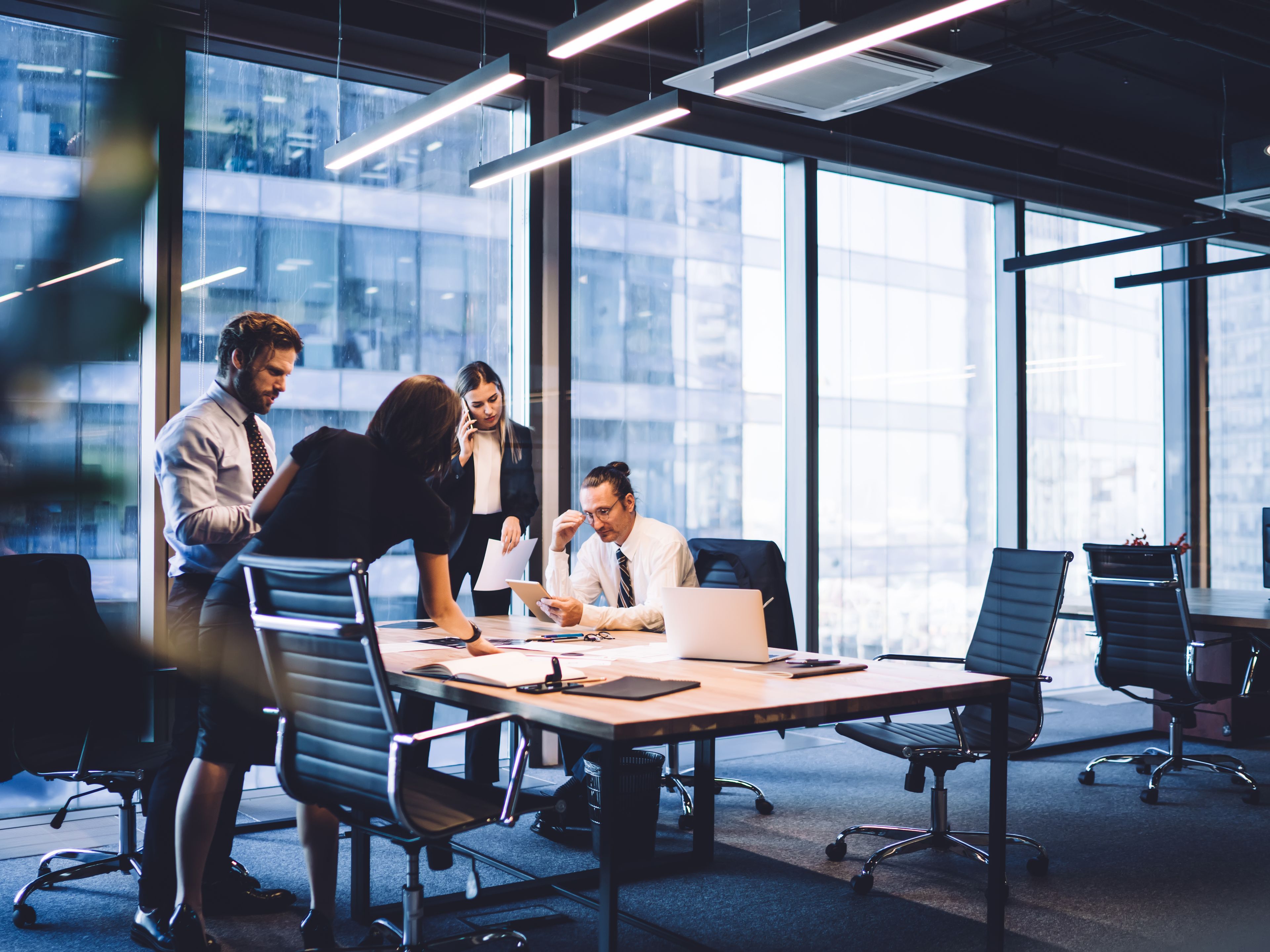 Sestanek štirih sodelavcev v sodobni pisarni v stolpnici z velikimi panoramskimi okni.
