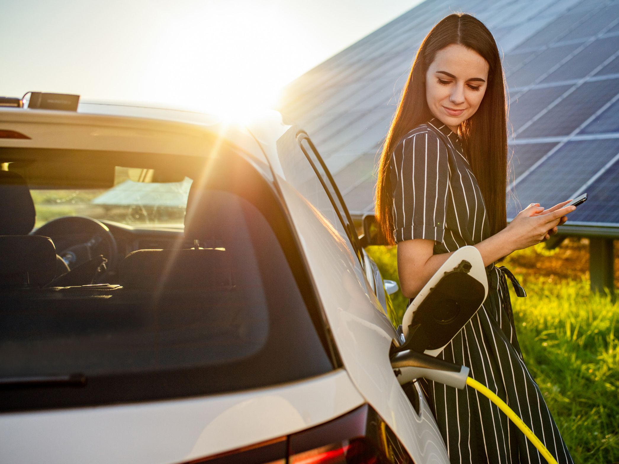 Ženska se naslanja na električni avtomobil, parkiran pred sončno elektrarno.