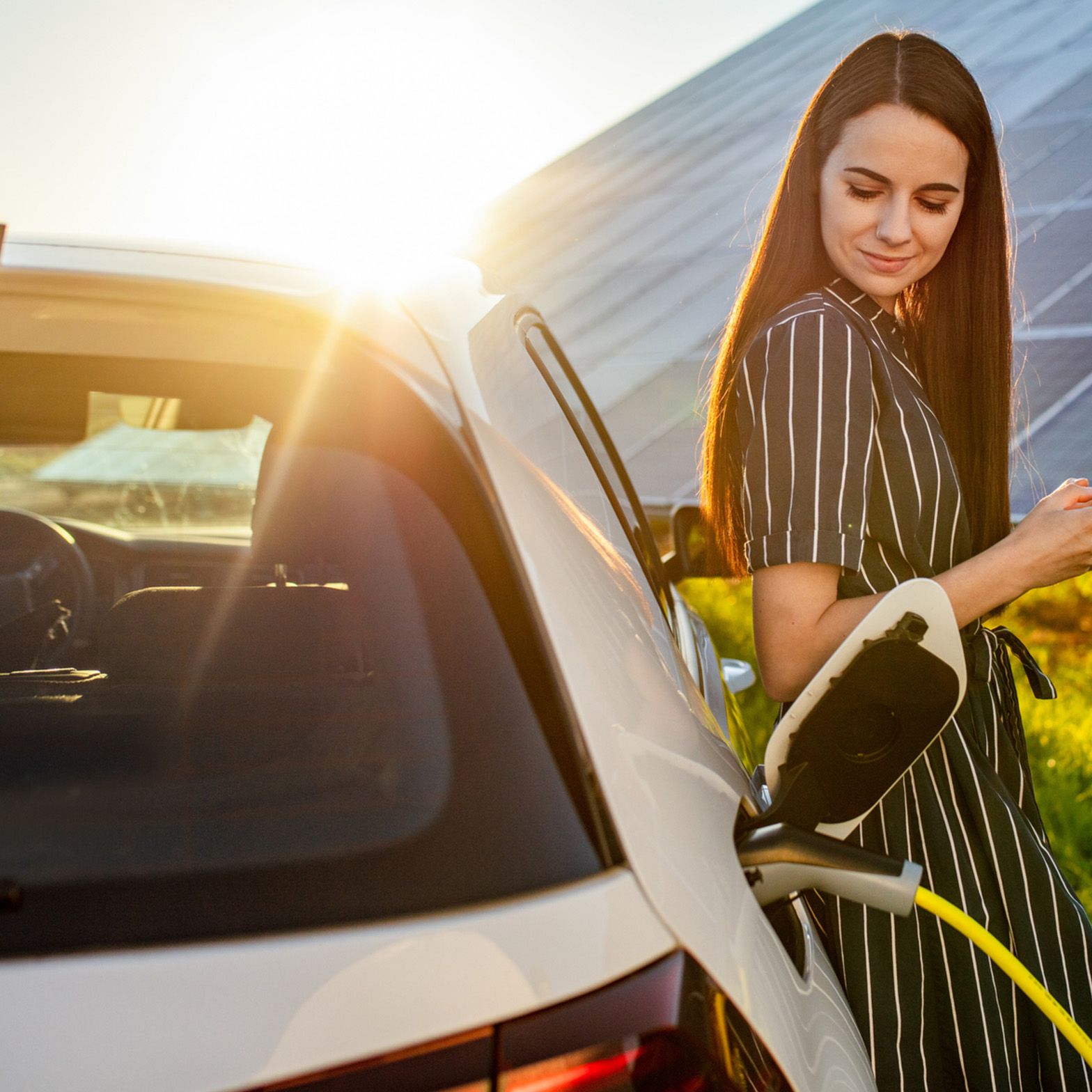 Ženska se naslanja na električni avtomobil, parkiran pred sončno elektrarno.