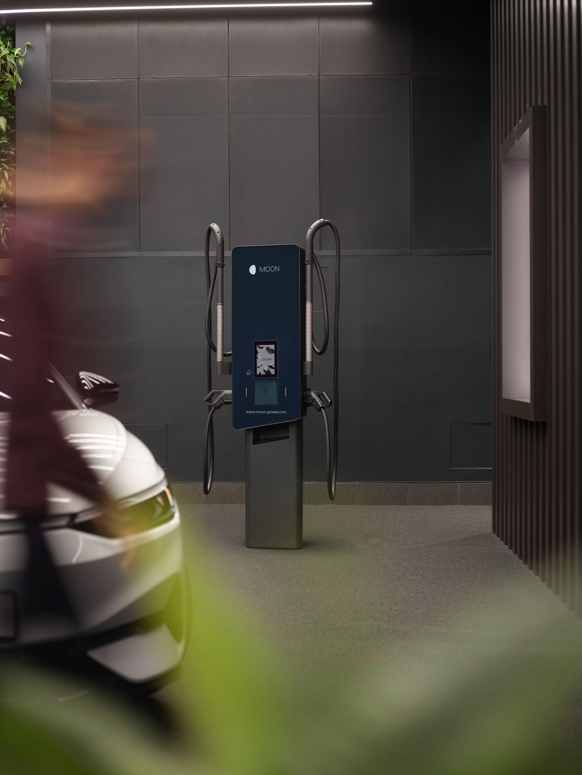 MOON stenska polnilnica v prostoru z sivimi stenami, v ospredju pa električni avtomobil.
