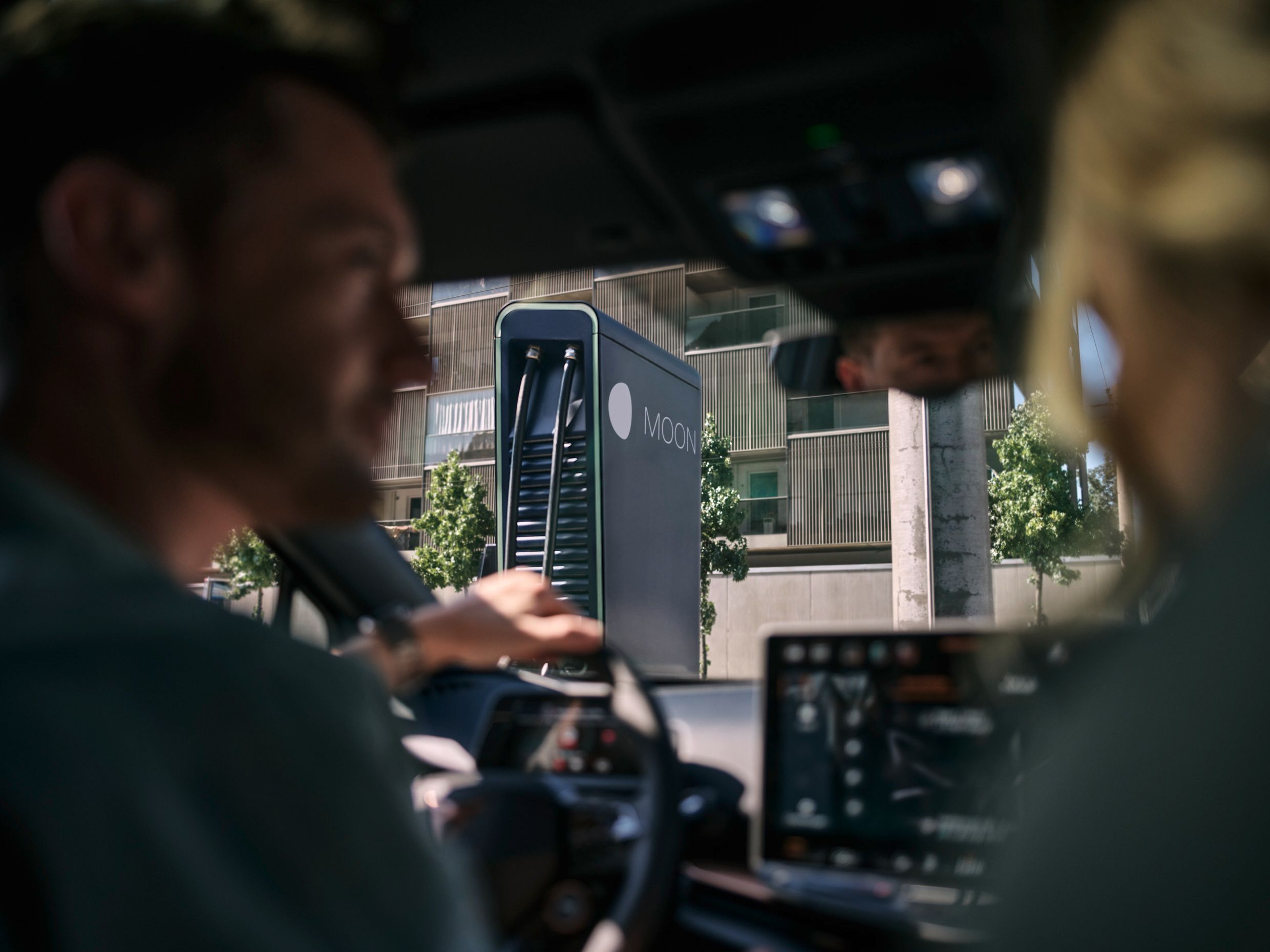 Dve osebi sedita v avtomobilu, parkiranem pred MOON hitro polnilno postajo.station