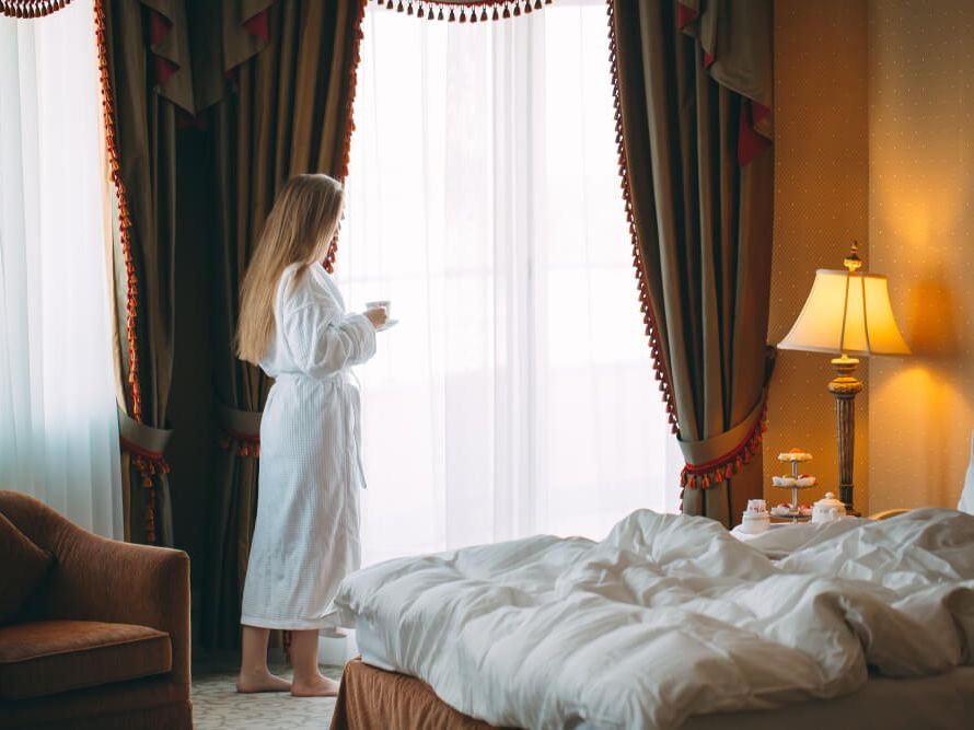 Ženska v hotelski sobi v kopalnem plašču stoji ob oknu in drži skodelico.
