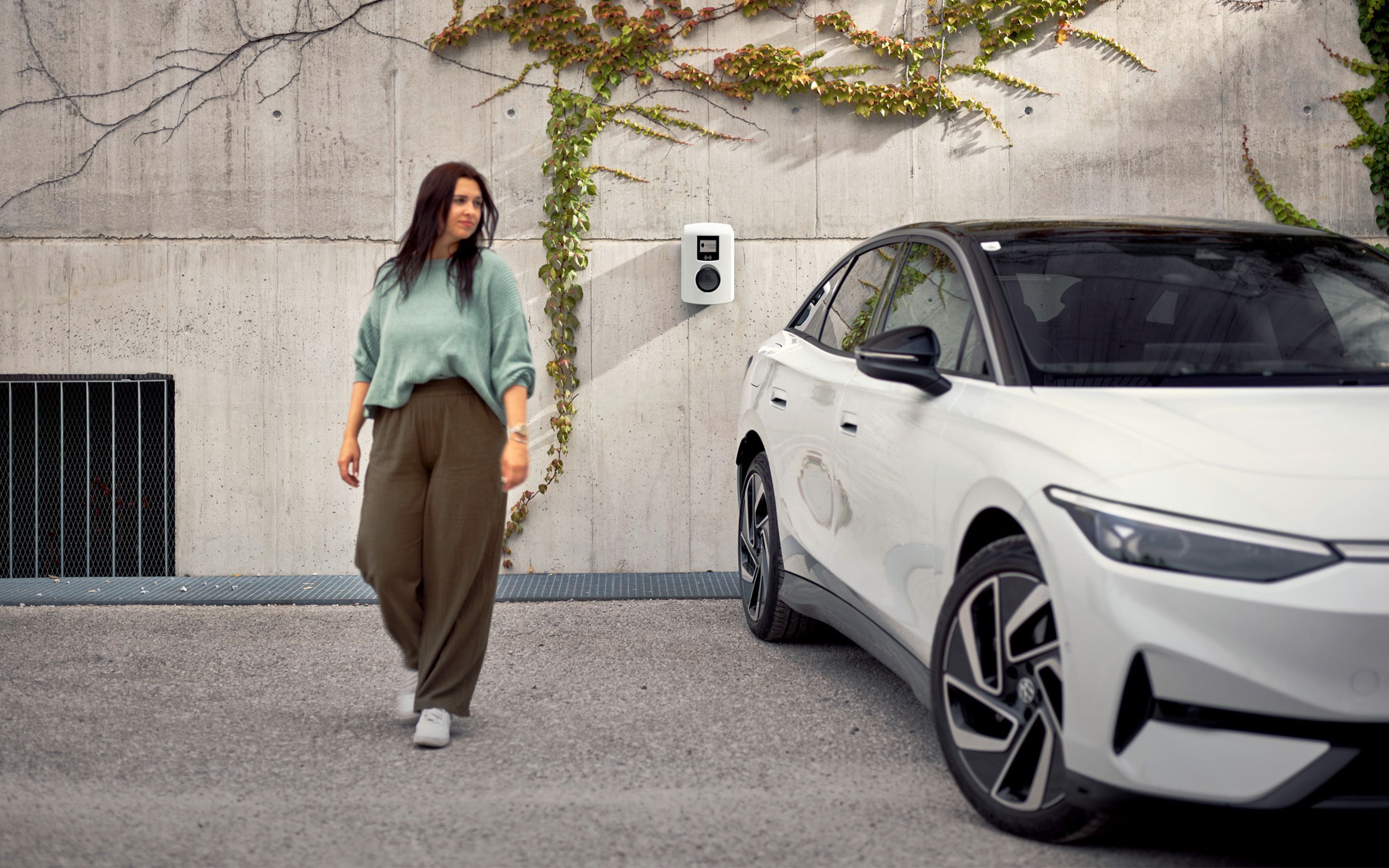 Ženska hodi mimo belega električnega vozila, parkiranega pred MOON Single Pro‑Line stensko polnilnico.